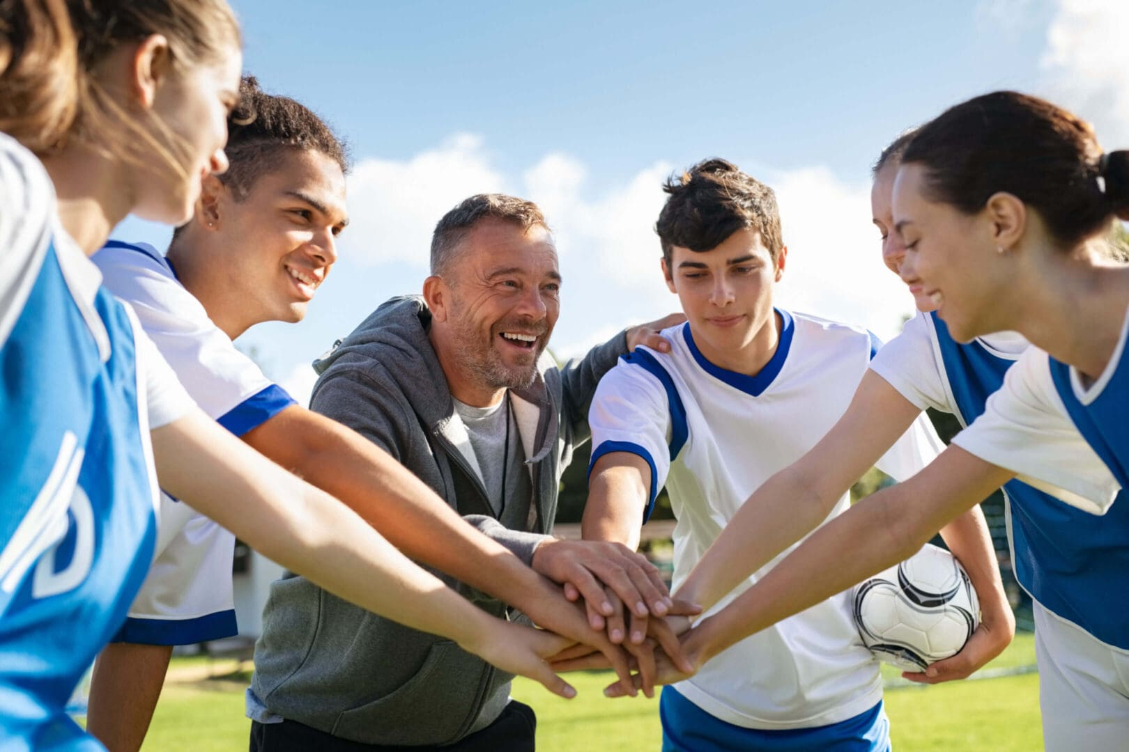 Coach and young soccer players join hands in a team huddle outdoors.
