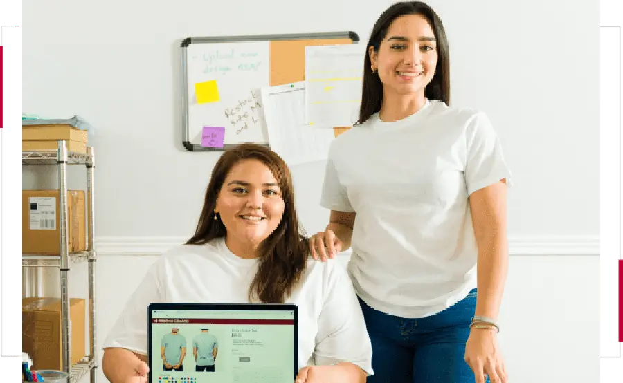 Two women in white shirts smiling, one seated with a laptop showing a website.
